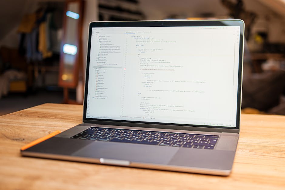 A close-up view of a silver laptop with a black keyboard placed on a wooden table in a well-lit indoor environment. The screen displays lines of code in a text editor, with a blurred background featur